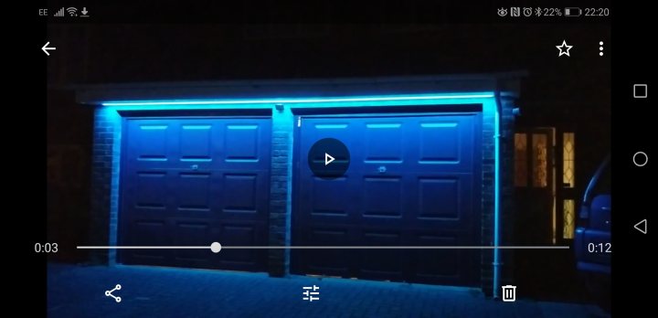 LED tape light for outside with timer? - Page 1 - Homes, Gardens and DIY - PistonHeads - The image shows a night scene featuring an illuminated garage door. The garage has a blue neon light along its length, creating a dramatic effect against the dark background. In the foreground, there is a smartphone with its screen displaying a video of the same garage door from a different angle. The phone is held in a landscape orientation, and the screen reflects the bright lights from the garage.