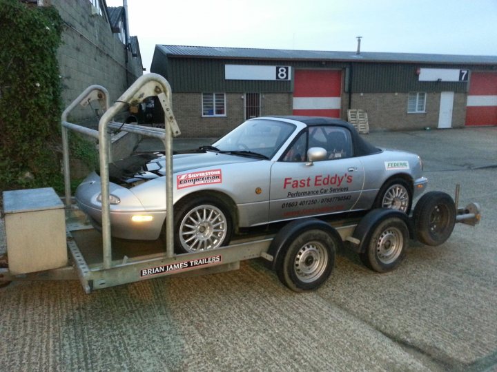 Looking forward to seeing everyone at Snetterton on Thursda - Page 1 - East Anglia - PistonHeads - The image shows a silver car on the back of a flatbed trailer, being transported by a tow truck. The car appears to be an older sports car, and is located next to a building with metal doors. A sign on the trailer indicates that the vehicle is being transported by "Brian James Trailers," suggesting that it might be a racing trailer. There are several numbers visible on the trailer, but the overall scene suggests someone is moving or transporting the car, possibly to or from an event or location.