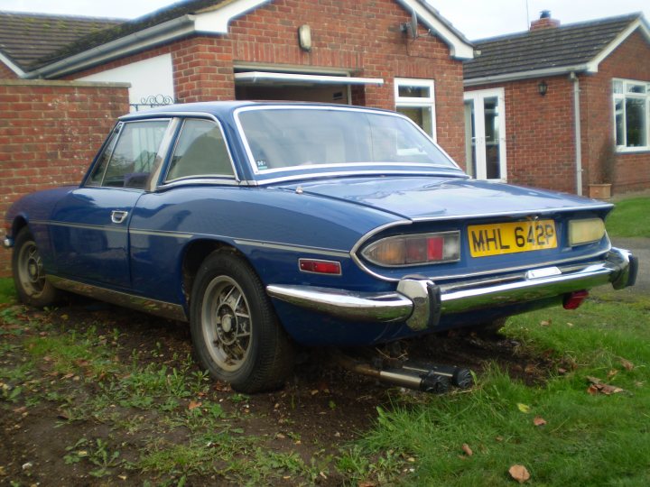 Scrap the Stagg, or give it a new home? - Page 1 - Triumph - PistonHeads - The image shows a blue MG car parked on a grassy area in front of a brick house. The car is an MG Midget, which is a classic English sports car known for its sporty handling and striking design. The Midget is parked facing the front of the house, with part of its bodywork visible. The vehicle appears to be in good condition and the surroundings suggest a domestic setting. The photo seems to be taken during the day, with the sky visible in the background, dominated by what looks like an overcast or cloudy day.