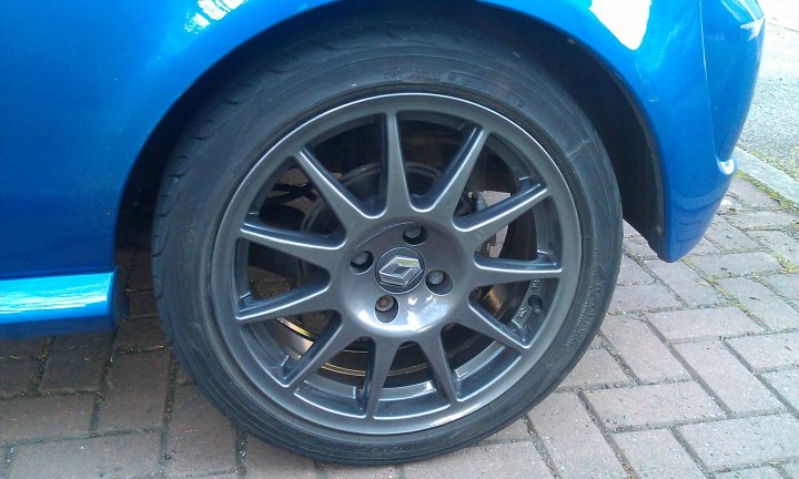 Alloy Wheel Refurb Aberdeen? - Page 1 - Scotland - PistonHeads - The image displays the side view of a blue car, focusing on its front wheel. The rim of the wheel is fashionable and silver, contrasting with the black tire. The patterned bricks of the road provide a textured backdrop to the scene. The car appears to be in good condition with no visible signs of damage or wear. The perspective of the photo is from a slightly lower angle, giving a more intimate view of the wheel and tire.