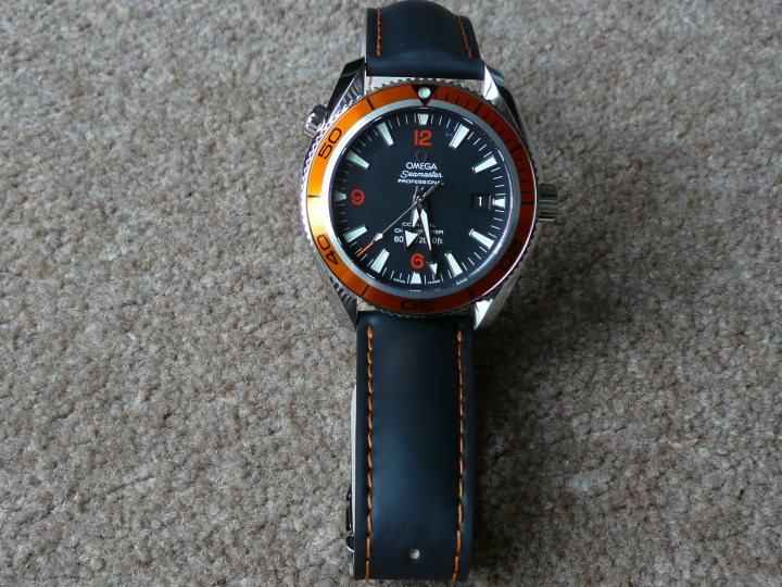 Pistonheads Seamaster Leather - The image displays a wristwatch with a black and orange case, featuring both interactive and analog controls. The watch has a black leatherstrap, and it is set against a textured, light-colored surface that resembles carpet. The minute hand of the watch points toward the number 8, which is one of the visible markers on the face. The watch brand is not distinctly displayed, but it appears to be a professional or luxury brand given the craftsmanship and quality of its design. The watch is depicted in a clear and detailed manner, showcasing its design and features.