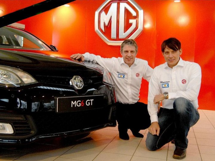 Are MG6's going to sell well? - Page 1 - MG - PistonHeads - The image displays two men in a showroom, posing with a black car positioned in front of them. The car has a large grille and prominently displays the badge "MG6 GT." Both men are wearing suits and each has a small badge on their shirt, which also features a logo, suggesting they may be representatives or owners associated with the car brand.