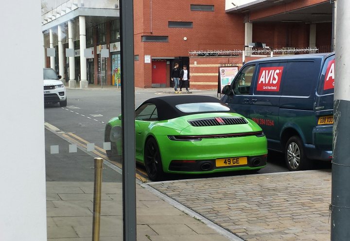 Spotted In South Wales (Vol 3) - Page 214 - South Wales - PistonHeads - The image is a photograph taken from the perspective of a person standing inside a building, looking out onto a street scene. In the foreground, a vibrant green sports car is parked along the curb, facing towards the viewer. Behind the car, there's a small commercial vehicle parked. A black van is also visible in the background. The street appears to be in an urban area with buildings and shops lining the sides of the road. The sky is overcast, suggesting it might be raining or have recently rained.