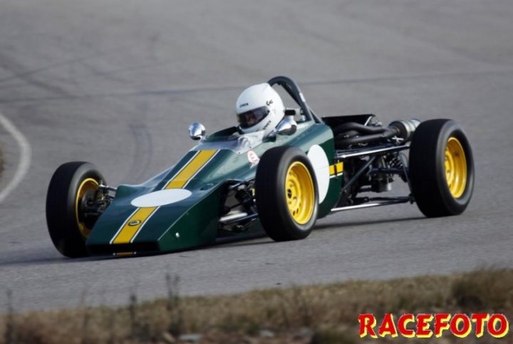 caterham racing colour code? - Page 1 - Caterham - PistonHeads - This is a dynamic racing photo featuring a single-seater race car with dominant green and yellow livery, positioned on what appears to be a race track. The car, angled to the left, is captured in a racing pose with significant lean into the turn. The driver's visor is lowered, and they appear focused on the track ahead. The surroundings suggest a daytime setting with clear visibility, scattering highlights across the picture, which indicates bright light conditions.