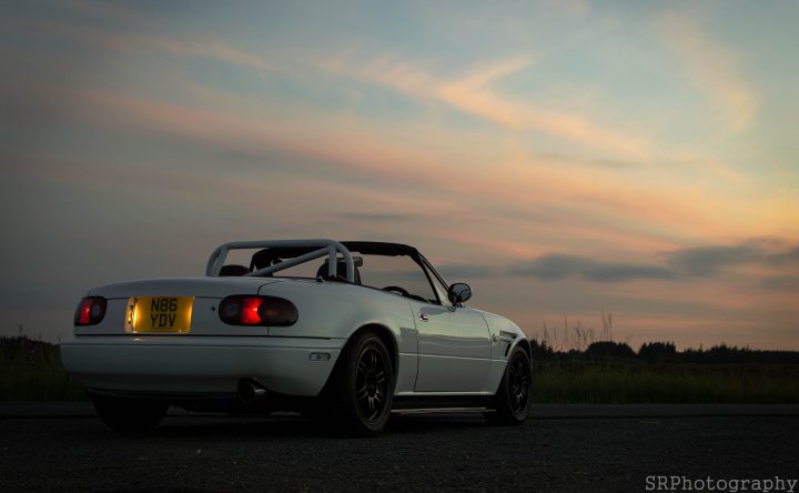 Our Cars - Page 136 - Scotland - PistonHeads - The image features a silver convertible car at dusk, with the sky transitioning from daylight to the colors of sunset. The car is parked on an asphalt road and is facing to the right of the frame. The pop-up roof of the convertible is down, and the car's headlights are shining, indicating the start of a journey or preparations for one. The scene conveys a sense of anticipation and the openness of road travel.