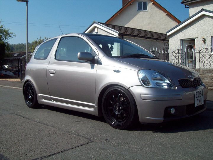 hi guys im new here...... - Page 1 - South Wales - PistonHeads - The image showcases a gray car parked on a residential street. The car is a hatchback model, with a two-door design featuring alloy wheels, which are distinctively black. The vehicle's body is chromed, adding a touch of elegance to its overall appearance. The setting is quiet and suburban, with houses visible in the background and a light pole nearby, suggesting the photo was taken during the day, under clear sky conditions.