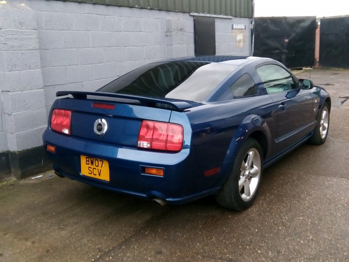 What to look out for - Page 1 - Mustangs - PistonHeads - The image shows a parked blue Ford Mustang with a black stripe on the hood. It is parked in a lot, possibly outside a building as suggested by the presence of a brick structure in the background. The car's license plate reads "SVY." There are no people visible in the picture. The lighting suggests it might be late afternoon or early evening.