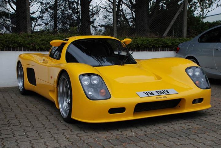 RE: PistonHeads Sunday Service: 25/07/10 - Page 9 - Events/Meetings/Travel - PistonHeads - The image features a unique, bright yellow sports car. The car is positioned on a cobblestone surface and it has a distinct design that includes side windows and headlights. The license plate reads "YB OHV," and there's a notable gap in the middle of the car's nameplate. The vehicle appears to be parked, with its rear facing the viewer, and it is contrasted by a grey and white car parked behind it. The scene suggests an outdoor setting, possibly a parking area or an event.