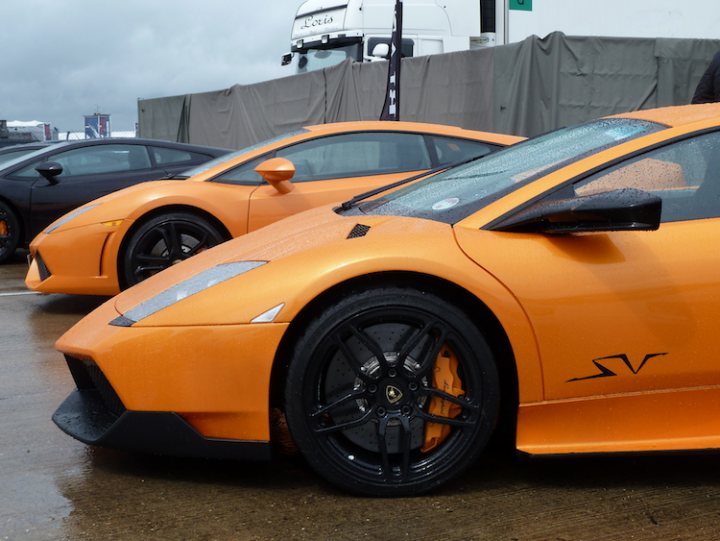 RE: Lamborghini Gallardo LP550-2: PH Carpool - Page 3 - General Gassing - PistonHeads - The image captures a collection of sports cars parked on a wet surface, reflecting their vibrant colors. Dominating the scene is a bright orange car, identical to the one beside it. The wet surface is crisscrossed with tire tracks, suggesting the activity of the cars. The background features a truck, contributing to the scenic setting.