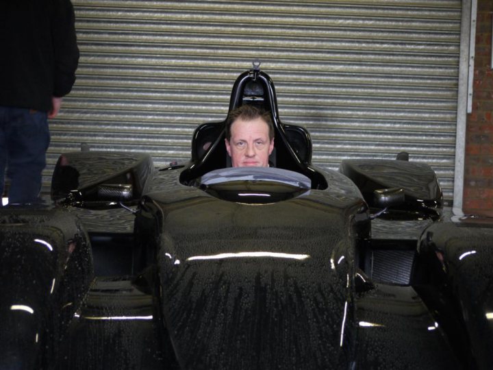 Snaps Secret Pistonheads - The image features a man sitting in a race-like car, which is positioned within a garage setting. He appears to be looking directly at the camera. His expression and the context suggest that this might be a shop or a workshop for racing vehicles. Another person, out of focus, is visible in the background. The car itself has a sleek, streamlined design, indicative of speed and performance.