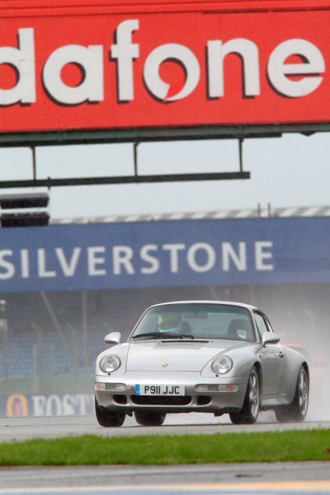 Pictures of your classic Porsches, past, present and future - Page 32 - Porsche Classics - PistonHeads - The image features a silver sports car with a Forza livery, navigating a wet racetrack. The car is in motion, leaning slightly to one side under the sodden conditions. The venue is clearly a professional racing circuit, indicated by the trackside sign reading "Silverstone." The overall atmosphere is one of excitement, as viewers anticipate the thrilling speed of the racecar.