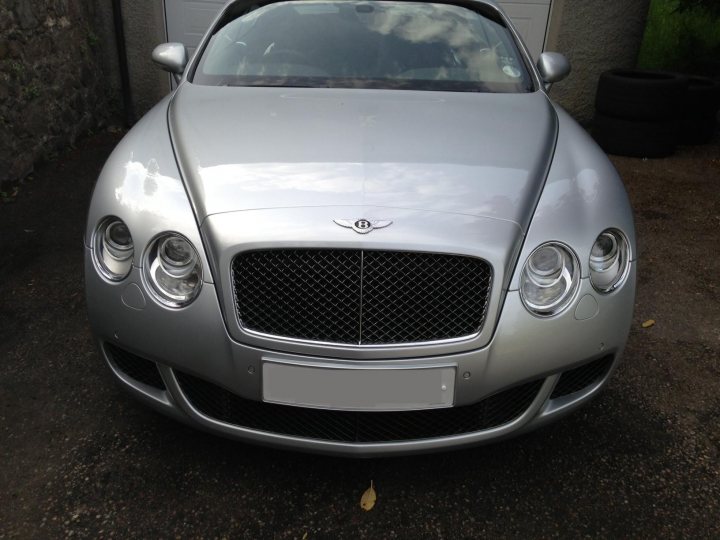 I’ve put my football boots on & bought a Bentley GT Speed - Page 1 - Rolls Royce & Bentley - PistonHeads - The image displays a silver-colored Bentley car. The vehicle is parked on a lot or driveway, with its sleek, curved silhouette dominating the foreground. The Bentley icon, a round emblem resembling a clock, is prominent in the grille. The car's construction suggests a level of luxury, and its polished finish indicates it is well-maintained. The setting is outdoors during daylight.