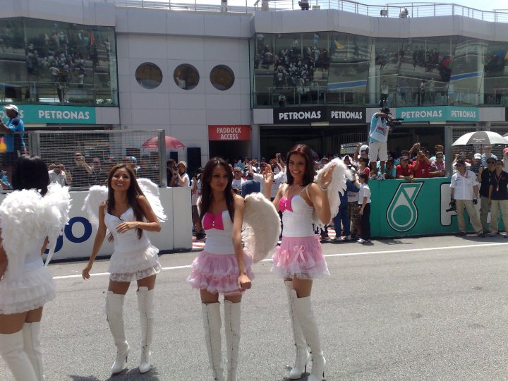 Afos Pistonheads Sepang - The image captures a lively scene on a city street. In the foreground, three women dressed as angels are standing and waving to the crowd. They're all wearing white dresses with wings, and one of them has a pink bow on her attire. Behind them is a building adorned with the word "Petronas" and a green leaf symbol. The street is lined with spectators, some under umbrellas and others holding cameras, all watching the angel-clad women with interest.