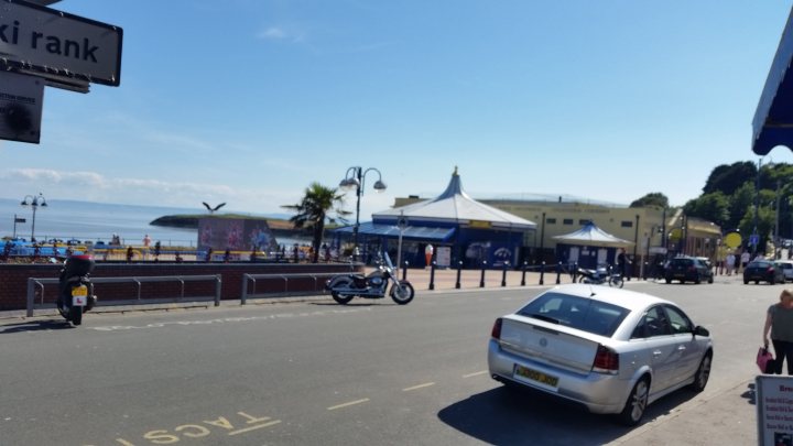 Pistonheads - The image captures a lively street scene on a sunny day. Parked cars and a motorcycle line the street, giving the impression of a bustling city life. Pedestrians walking on the sidewalk add a sense of liveliness to the scene, while a bus signifying a bus stop indicates the availability of public transport. A waterfront is also visible in the background, contributing to the overall urban atmosphere. The sky is a clear blue, suggesting good weather conditions.