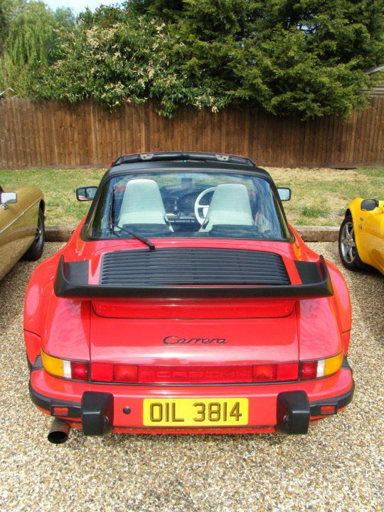 Breakfast Meeting, The Hare, Roxwell 4th June - Page 1 - Kent & Essex - PistonHeads - The image showcases a vibrant red Porsche parked on a gravel driveway. The car is a 911 model, identifiable by its distinctive black rear wing and a license plate that reads "OIL 3811". The Porsche's design and features, along with its sports car identity, suggest it's likely well-maintained and frequently driven. The backdrop of a wooden fence and trees contribute to a picturesque, suburban setting.