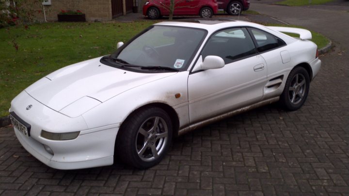 MR2 specialist near Sherborne? - Page 1 - South West - PistonHeads - The image shows a white two-door coupe parked on an asphalt driveway. The car has a sleek, aerodynamic design with a hood vent and a large rear spoiler. Its alloy wheels and lowered suspension suggest it may be a modified or tuned vehicle used for speed or style. In the background, there is a suburban house with a red car and a partly cloudy sky above. The image is taken during the day under natural lighting conditions.