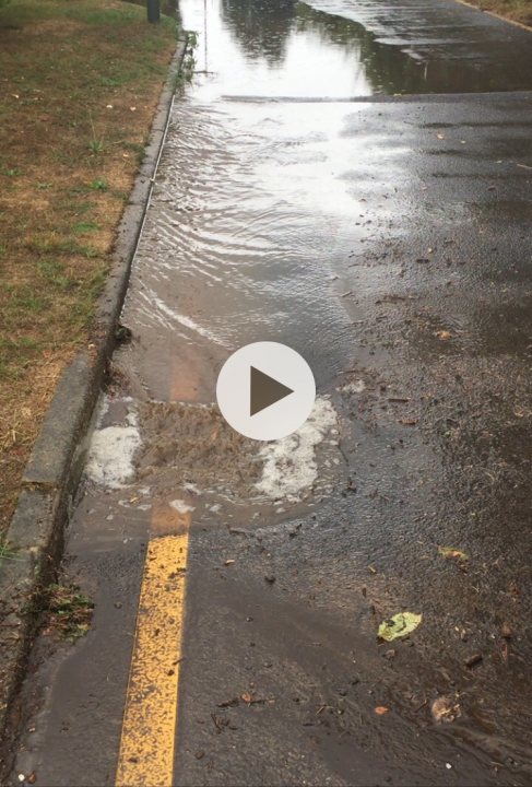 House buying - Seller states No to flooding - Page 3 - Homes, Gardens and DIY - PistonHeads - The image shows a flooded street with water reaching almost to the top of the road. This scene is captured on a smartphone screen, as indicated by the visible interface elements such as a battery indicator and a camera icon in the corner. A small portion of the video paused frame can be seen, showing the water level extending from the road onto the sidewalk. The water appears brown, possibly due to the presence of mud or sediment. In the background, there are trees, suggesting that this street is located near a wooded area. The overall impression is of heavy rainfall leading to localized flooding.