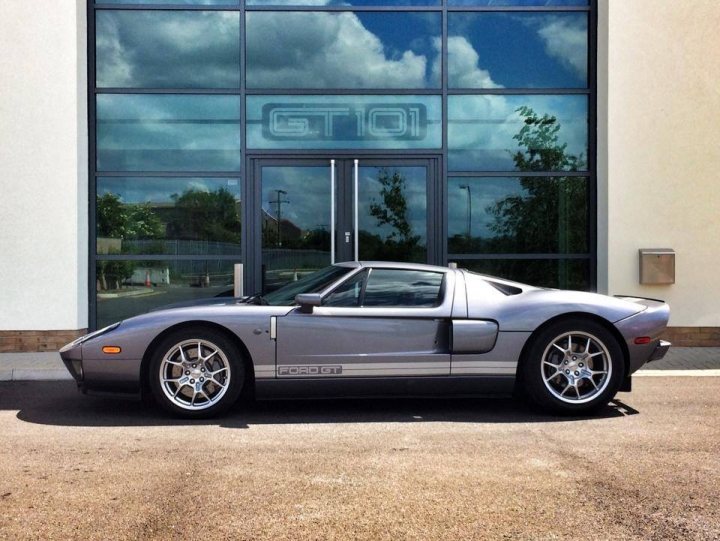 Just bought an old Ford - Page 1 - East Anglia - PistonHeads - The image shows a silver, two-door sports car parked in front of a large building with a distinctive glass facade that reflects the image of the car. The reflection is clear and symmetrical, creating a mirror image of the car on the building's surface. The car is positioned where it appears to be driving towards the camera, giving the impression of movement. The background is mostly out of focus, but the car and the reflective building are the main subjects of the photograph. The car's sleek design and the bright and possibly sunny conditions provide a dynamic feeling to the image.