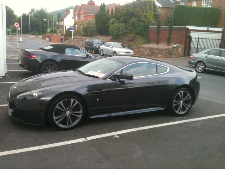 Hi from a newbie - Page 1 - Aston Martin - PistonHeads - In the image, a black Aston Martin DB7 car is the main subject, parked in a parking space within a lot that is marked with white lines. The car is facing the camera, and a pair of redeemers are attached to the windshield. Behind the DB7, two other cars are visible in the background, one a silver sedan and the other a black crossover. The setting appears to be an urban area with houses and businesses in the distance. The sky is overcast, suggesting a cloudy day.