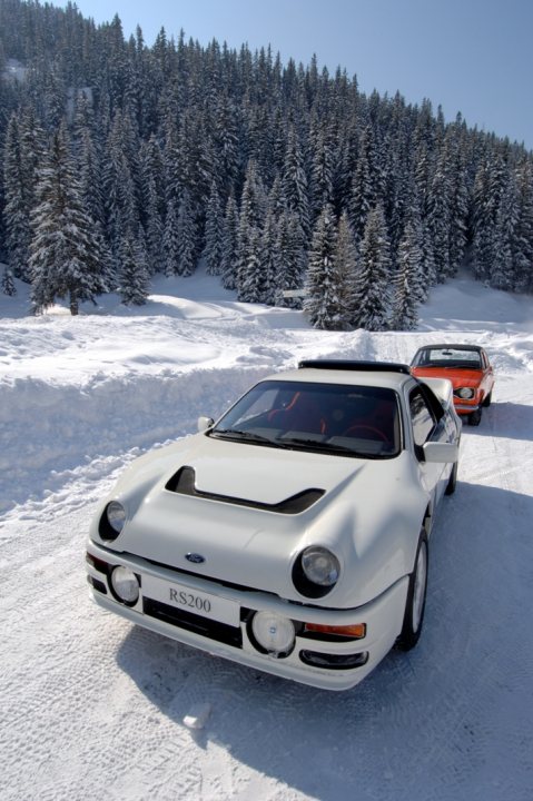 Pistonheads Scenes - The image captures a serene winter scene with two vehicles prominently displayed. The foreground features a white, racing-style sports car, presumably a RS200 model, resting on a snow-covered road. This car is adjacent to a red pickup truck. Both vehicles cast shadows on the snow, indicating they are in the sunlight. The backdrop is a picturesque landscape of snow-dusted trees and a clear blue sky. The overall scene suggests a well-maintained road cutting through a beautiful, snowy forest.