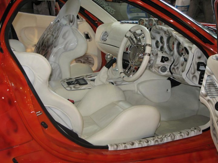 Sydney Pistonheads Autosalon - The image features the interior of a car with a two-tone color scheme, where the upper part is white and the lower part is orange. The car is equipped with multiple controls, including a steering wheel and pedals on the floor. There are also various buttons and dials on a control panel, as well as a center console that likely houses additional controls. The side door is partly visible, showcasing some of the design and materials used in the vehicle's construction.