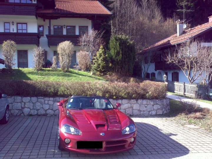 Lovley Pistonheads Day - In the image, there's a vibrant scene featuring a red sports car parked by a brick driveway. The sports car is sleek and shiny, with a prominent grille and headlights that suggest speed and power, reminiscent of a racing look. The driveway is adjacent to a house with a well-maintained garden, offering a serene backdrop. In the foreground, there's a paved walkway that seems to be recently cleaned, leading the viewer's eye towards the house. The car is positioned to face the viewer, displaying its front view, which draws attention to its stylish design and colors. The overall setting seems to be a residential area with lush trees in the background, contributing to the tranquil ambiance.