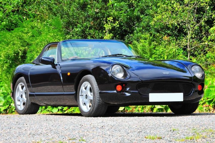 Chimaera Pistonheads - The image shows a black convertible sports car parked outdoors. The vehicle has its top down, allowing the driver to enjoy the open air. The car features two doors, typical of a two-door sports car model. The car is situated on a gravel driveway in front of a lush green bush. The lighting and shadows indicate that the photo was taken during the day under clear skies.