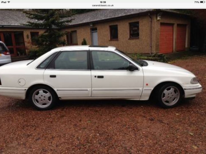 Classic (old, retro) cars for sale £0-5k - Page 133 - General Gassing - PistonHeads - The image shows a white sedan parked outdoors on a gravel driveway in front of a house. The car has four doors and alloy wheels. There are residential buildings in the background with red doors, one of which appears to be an open wooden garage door. The wall of the house is red, complementing the natural red color of the gravel. The photo seems to have been taken during the day under cloudy skies. The car is of older model, as indicated by its design.