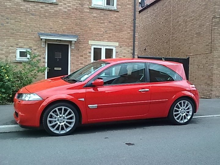 Replacement Pistonheads - The image features a vibrant red sedan parked on the side of a street. The car is positioned next to a house, allowing for a clear view of the brick wall and the door of the house. The car's side mirrors are reflecting the clear blue sky above, suggesting a bright, sunny day. The wheel rims of the car stand out against the red body, adding a fresh-painted feel to the scene.