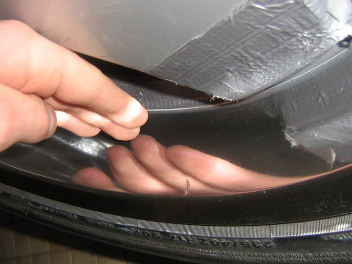 The image features a hand reaching in towards a tire, which has some tinfoil or similar material on its sidewall. The fingers of the hand are seen touching the black and grey rubber of the tire. The tire has some visible use; it shows signs of wear and marks that suggest it's an older, possibly well-used tire. The atmosphere of the image is one of close inspection, possibly for maintenance or inspection purposes.