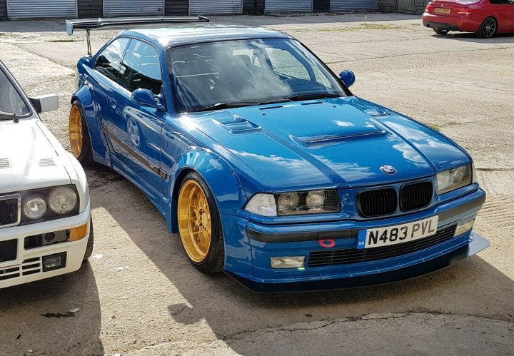 Pictures of decently Modified cars [Vol. 2] - Page 1 - General Gassing - PistonHeads - The image shows a parking lot with two vehicles. The prominent car is a blue BMW, which appears to be customized with gold wheels and a large spoiler at the back. This car is parked next to a white BMW. In the background, there are other cars visible and a building. The setting suggests an outdoor location, possibly a public parking space or a dealership lot.