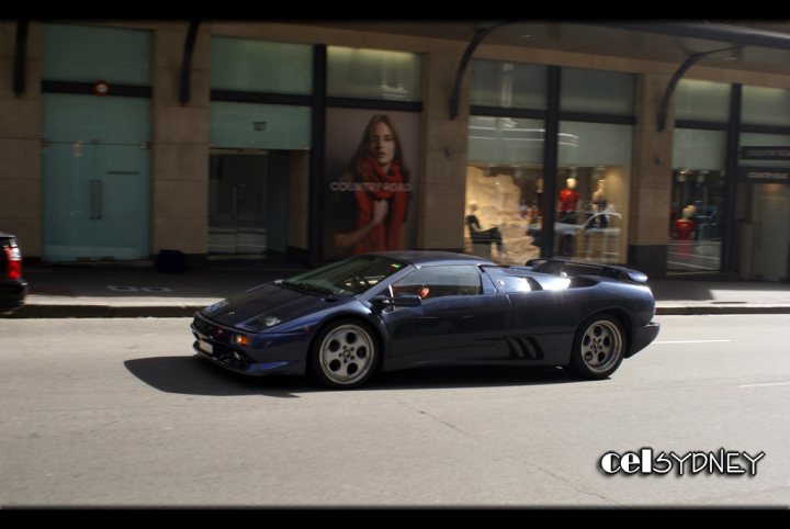 Show us your Diablo  - Page 6 - Diablo/Murcielago/Aventador - PistonHeads - The image captures a moment on a city street where a vintage sports car, painted in a sleek blue, is the main focus. The car, parked on the side of the road, flawlessly reflects the light from above, enhancing its glossy appearance. Across the street, a contemporary building stands, punctuated by a window display featuring numerous stylish mannequins. The contrast between the classic sports car and the modern fashion boutique adds a sense of timeless elegance to the scene.