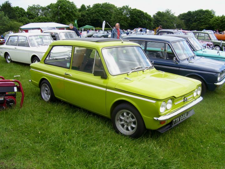 Lookalike Pistonheads Minilites - The image shows a gathering of vintage cars on a grassy lawn. The most prominent vehicle is a bright green two-door car with distinctive design lines. This car is parked in front of a white car which has a sunroof and is slightly obscured by the green vehicle. Behind the white car, there are additional vintage cars, some of blue and white, while others are a mix of colors. The setting suggests an outdoor event or car club meetup, with people walking around the vehicles.