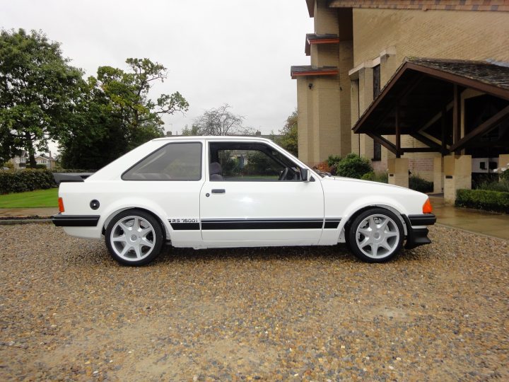 Escort RS1900i?? - Page 3 - General Gassing - PistonHeads - The image shows a white sports car parked outdoors on a gravel driveway. The car has a clean and sleek appearance with black and white stripes along its body. It is a two-door model with a distinctively wide stance, large black trim, and five-spoke alloy wheels. The car is stationary and appears to be a German or European production vehicle. The background features a building and some trees, suggesting a residential area.