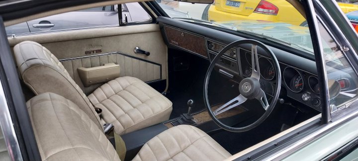 Pistonheads - The image displays an old-fashioned car with a cream interior and black seats, parked indoors. The car features classic design elements such as chrome trim around the windows and body, a prominent steering wheel, and a dashboard with multiple gauges and controls. A yellow object is visible in the background behind the vehicle, but its nature isn't clear from this angle. The photo appears to be taken from an elevated perspective, possibly looking down at the car, providing a view of the seats and interior details.