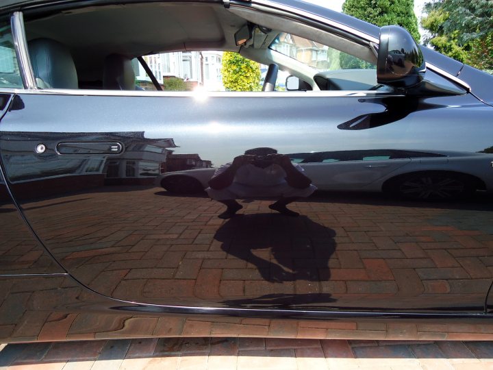 Pistonheads Detailed - The image presents a clear view of a person, as seen reflected in the shiny surface of a car on a brick parking lot. The person seems to be in a crouched or low position, as their body is partially obscured by the vehicle. The car itself appears to be black and sleek, with the reflection showing a slight sheen on its surface. In the background, there are other cars visible, suggesting this could be a public parking area. The ground is covered with red brick pavers, providing a neutral canvas for the scene.