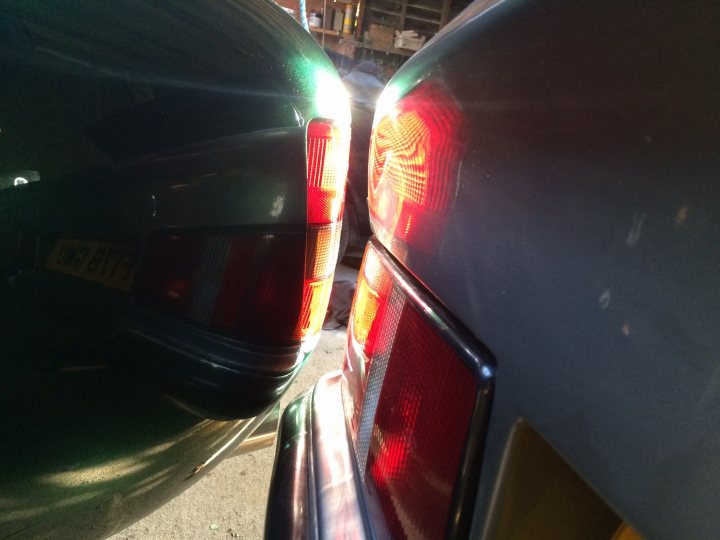 Random TVR pics - Page 409 - General TVR Stuff & Gossip - PistonHeads - The image shows a close-up perspective of two vehicles, with a focus on the left side rear light area. The car appears to be green in color, while the other vehicle is white and metallic. The reflection of sunlight on the car surfaces suggests the time is midday. The details visible include the brake light, turn signal, and rear wiper, indicating this is the back of the vehicles' bumpers. The angle of the shot is slightly upward, between the two cars, which gives depth to the image.