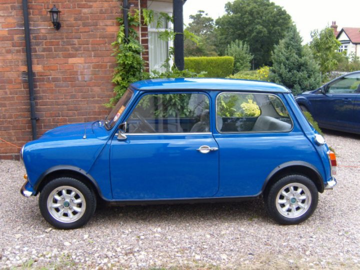 My Classic Mini 1000 - Page 1 - Readers' Cars - PistonHeads - The image depicts a charming blue mini classic car parked alongside a flagstone driveway. The car has a distinctive front end with round headlights and a small rear window. A black door handle suggests classic design elements, and the overall appearance of the car, including its color and style, gives it a nostalgic look. The vehicle's location on the driveway contrasts nicely with a modern blue car visible in the background, adding an interesting comparison between older and more contemporary vehicle styles.