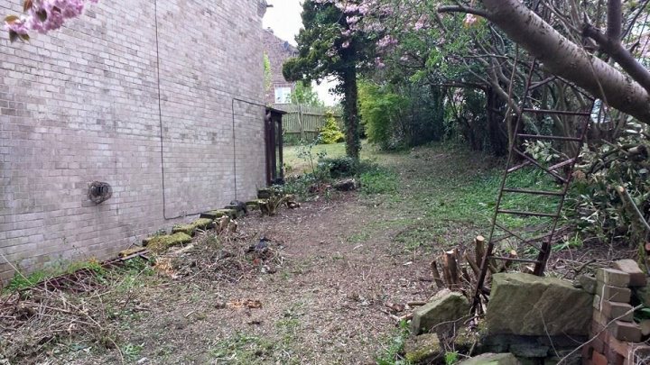 2015 Lawn thread - Page 22 - Homes, Gardens and DIY - PistonHeads - The image shows a narrow, uneven footpath leading through what appears to be a garden area adjacent to a building. The path is bordered by a mix of vegetation, including a bush with purple flowers near the entrance to the path in the foreground. A small, derelict gate is visible in the middle of the path, and the surroundings include bare plants and fallen branches. The construction and vegetation suggest the garden is not well-maintained. The sky is overcast, casting a soft, ambient light over the scene.