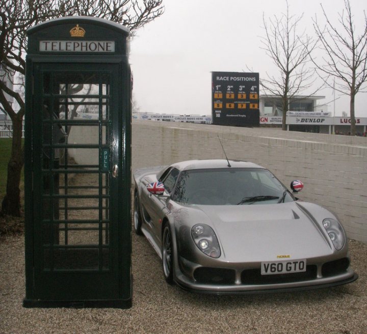 A car parked next to a parking meter - Pistonheads - This image depicts a vintage sports car and a quirky green telephone booth as the main subjects. The car, a sleek silver model, is parked on what appears to be a gravel or dirt lot, adjacent to a textured white concrete wall. The phone booth, topped with small white doors and a sign reading "TELEPHONE," stands by the roadside within the frame of the image. The setting is outdoors, possibly in a tourist area, as indicated by the presence of a banner with text and logos in the background. The atmosphere suggests a blend of leisure and functionality.