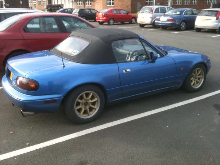 £3k, too steep? plus introduction - Page 1 - Mazda MX5/Eunos/Miata - PistonHeads - The image presents a serene parking lot scene featuring a vibrant blue two-door sports car. The sports car, which takes up most of the frame, has a sleek, hard-top design. The front end of the car is particularly eye-catching with its customized yellow and black wheels, and it is driving on the pavement in the lot. The car is not alone; there are other cars parked neatly around it, demonstrating neat organization in the space. The parking lot itself is situated in front of a collection of buildings, suggesting this could be a commercial or residential area.