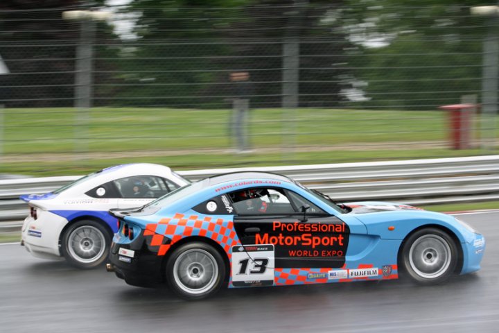 MSVR racing club champs at Brands - Page 1 - UK Club Motorsport - PistonHeads - The image captures a dynamic scene on a race track. Two cars are prominently featured in the image. The first car, painted in the iconic colors of the UK's lead racing team during the 2000s, is a blue and white checkered design. The second car is a bright blue with the text "Professional Formula World Expos" indicating that it might be associated with a world-wide tour of motor sport events. The cars appear to be in motion, possibly at high speeds, adding a sense of excitement and competition to the image. The background provides the context of the location, with a fence, suggesting this is a professional racing track.