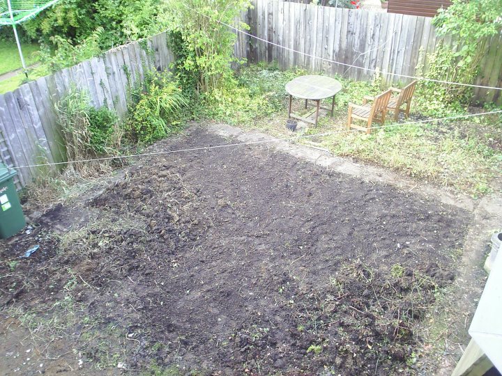 Pistonheads - The image shows a backyard with a garden area that is not yet planted. A garden plot is outlined with a wooden edge and is covered in soil, small plants, and mulch. There is a patio table and a chair placed on a concrete area to the left of the garden. A black trash can is leaned against the bottom left corner of a wooden fence. The yard is fenced in and appears to be overgrown with vegetation and a viny plant growing along the top of the fence. There are no visible buildings or structures in the image. The overall impression is of an outdoor space that is in a state of disarray and preparation for gardening.