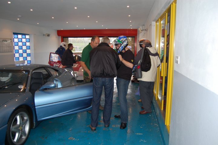 Pistonheads - The image captures a lively scene at a car shop. A group of people are gathered around a car, which is on a blue ground inside a workshop garage. The car, painted in a striking shade of blue, is the center of attention, attracting customers. 

Among the staff at the car shop, there's a man standing in the doorway with a purple shirt. On the other side of the blue ground, two more staff members are present - one of them is wearing a green shirt while the other is sporting a blue shirt. 

Additionally, another person is seen carrying a backpack, possibly a customer or a visiting salesperson. The workshop garage is well-lit and spacious, accommodating the car and the people comfortably. It's a typical day at the car shop, where people come together to discuss and admire the vehicles on display.