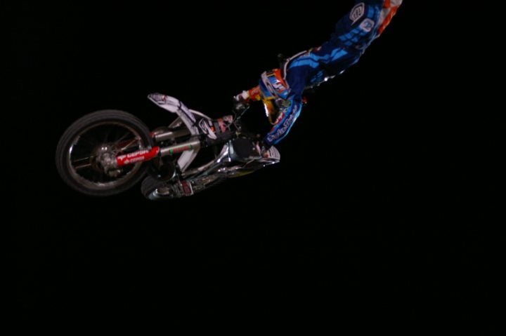 Bull Pistonheads Fighters Red - The image showcases a daring scene of a motocross rider in action during a nighttime event. The rider is performing an impressive jump, launching into the air atop a dirt bike. The bike's front wheel is elevated and angled towards the sky, while the back wheel has left the ground. The rider's body leans back and towards the right side of the image, indicating a powerful jump with proper form. The background is a blank, dark space, emphasizing the action of the rider and the contrasting between the illuminated dirt bike and the surrounding darkness.