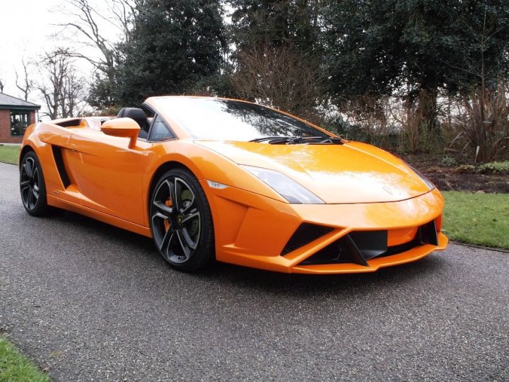 New arrival - Page 1 - Gallardo/Huracan - PistonHeads - The image depicts an orange sports car parked on a paved road. The car is sleek with a wide, flat front face and a noticeable elongated hood. It is positioned at an angle from which the rear wheels are visible. Behind the car, there is a grassy area with some plants, and a building can be seen further away on the left side. The sky suggests it might be cloudy.