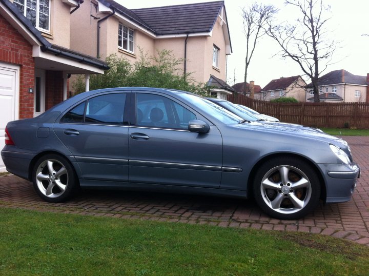 W203 Mercedes - as bad as they say? - Page 1 - General Gassing - PistonHeads - The image features a blue sedan parked on a grassy driveway. The car is a Mercedes Benz, identifiable by its sleek design and distinctive grille. The vehicle is positioned sideways to the camera, allowing a clear view of both the front and side profiles. The setting is residential, with a house in the background, and a well-maintained lawn and pathway under the car. The sky above is overcast, casting a soft light on the scene.