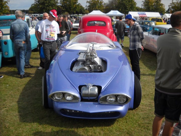 Anyone going to the NASC 40th Nats at Trinity Park? - Page 1 - East Anglia - PistonHeads - The image shows a car show where several vintage cars are on display. In the foreground, there's a striking blue and silver car with a prominent engine cover that is partially open. The engine appears to be a V8 or V12, and there's visible chrome on the body of the car. Surrounding the blue car are pedestrians, some standing and observing, while others are walking around due to the lively atmosphere of the event. The background consists of other colorful vintage cars, all with their unique designs and colors, adding to the vibrant and nostalgic ambiance of the show.