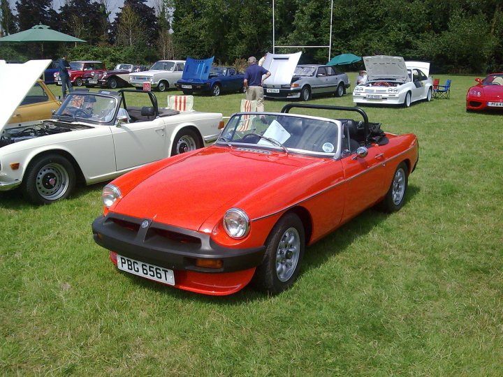 Classics Southern Worthing August Pistonheads - The image depicts a vintage car show set in a green field. The focus is on an old, restored red sports car with a black roof and a herringbone grille. It's positioned next to a pristine white convertible, both cars are on display and showcase their classic designs. The field is populated with other vintage cars parked under various umbrellas and tents, creating a vibrant and sunny atmosphere. The overall ambiance suggests this is a casual outdoor event dedicated to automotive nostalgia and beauty.