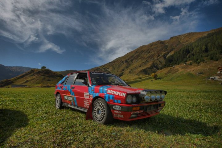 Some half decent pics - Page 1 - North West - PistonHeads - The image showcases a vintage race car parked in the middle of a vibrant and verdant grass field. The car is predominantly red with blue and black stripes running along its top and bottom. The car is equipped with four doors, two for the driver and two for the passenger, and features a large spoiler on the back. Beyond the car, the expanse of the grassy field is dotted with trees and mountains in the background, suggesting a remote and scenic location, perhaps on a tour of the Alps. The image captures the car in the daytime under a clear sky.